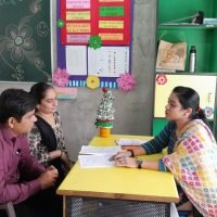 Teacher interacting with parents sitting across a desk in a decorated classroom during a meeting.