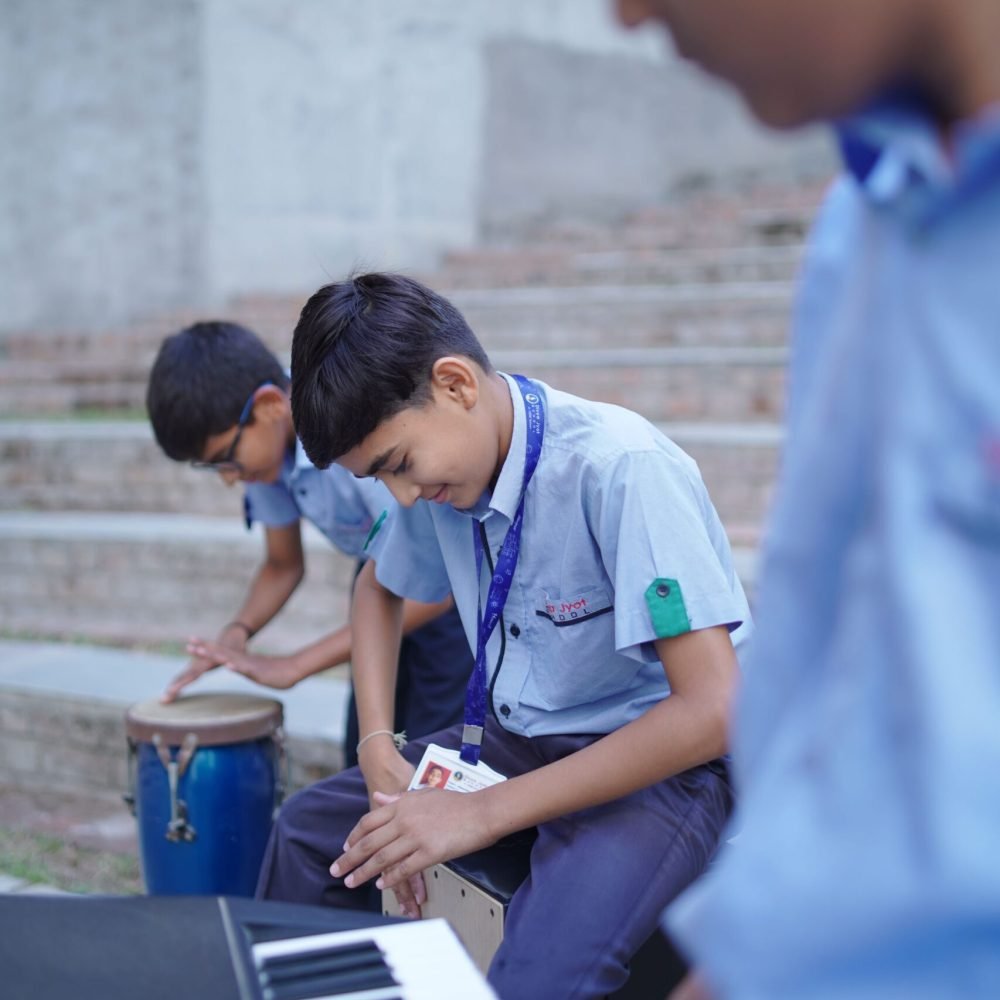 School students playing musical instruments outdoors, including a cajón drum, bongo, and electronic keyboard.