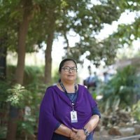 Female teacher in purple and blue traditional dress standing outdoors surrounded by trees.